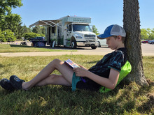 A boy reading a book leaning against a tree