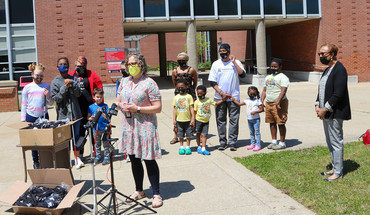 A woman speaks in front of microphones outside of a school building. Several students and other attendees stand behind her.