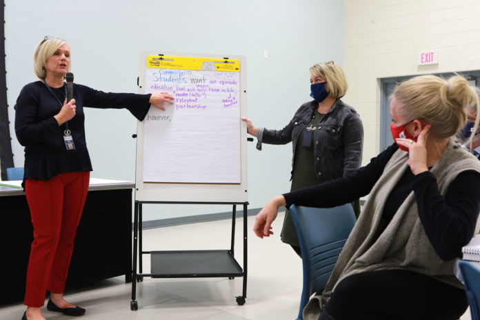 Picture of two women standing by a poster board with writing on it while talking to people in a large room.