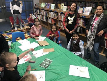 A group of parents and students gathered around a table with crafts on it. There are books on shelves behind them.