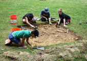 A picture of four students sitting on the ground, excavating a small square of ground using shovels and brushes.