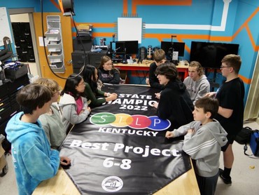 Students sit around a banner on a table that reads: State Champions 2022 STLP Kentucky, Best Project, 6-8
