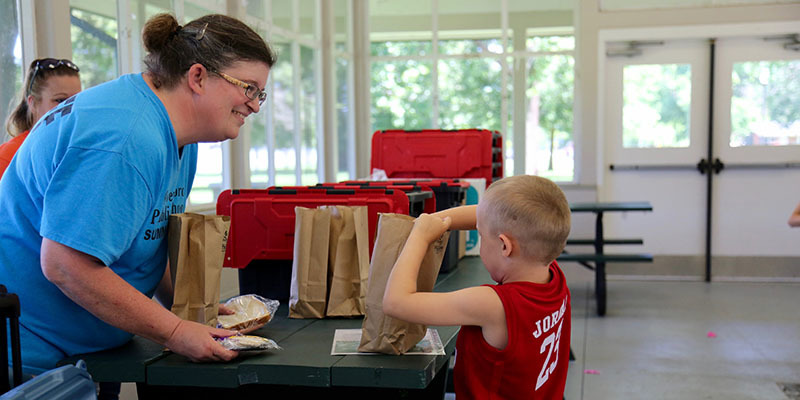 Picture of a young boy standing at a table, reaching into a brown bag while a woman smiles at him.