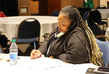 Picture of a woman sitting at a table in a conference room, writing in a notebook.