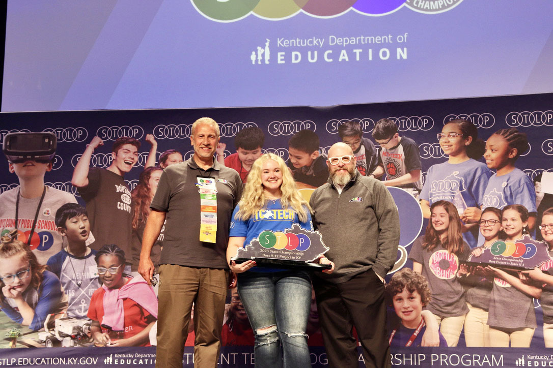 Picture of a student on stage holding an STLP trophy standing beside two Kentucky Department of Education staff members.