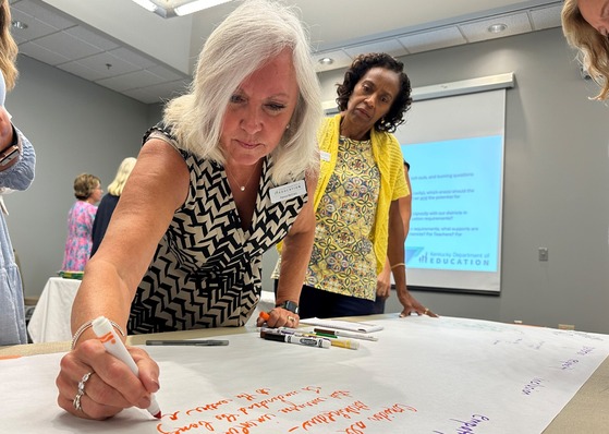 A woman is standing and using a marker to write on a piece of paper. Another woman looks on. Both are members of the Kentucky Board of Education.