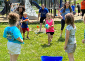 A group of kids run outside. A small girl with a sponge runs toward a bucket.