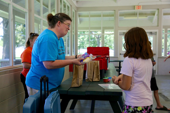 A woman hands a girl a lunch bag while smiling at her.