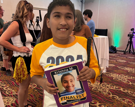 Krishna Malhotra stands in a large room holding a sign with his picture and the words 2024 Finalist, sophomore on it.