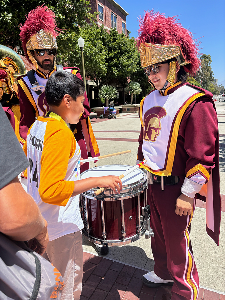 Krishna Malhotra stands outside in front of a University of Southern California drummer, holding onto drumsticks and getting ready to play the drum.