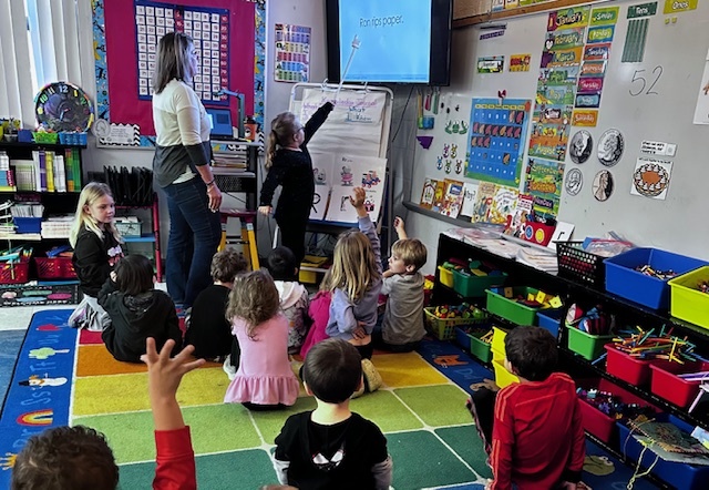 Student using pointer to read sentence off board while teacher and other students watch