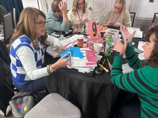 Five adults collaborate at a round table with colorful paper shapes, notebooks, and a table marker labeled “9.”