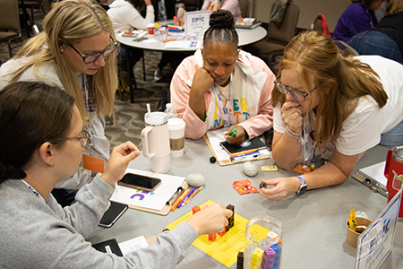 Adults seated at round tables collaborate using colorful math manipulatives during a workshop, discussing materials and recording notes