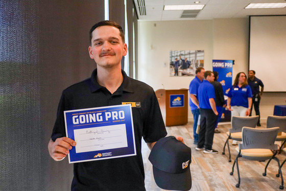 A student holds a sign that reads “Going Pro” while holding a black hat with the Caterpillar log