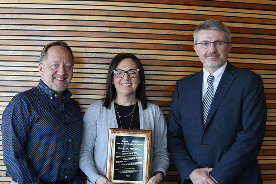 Three people pose for a photo in front of a wood-paneled wall. A woman in the middle is holding an award