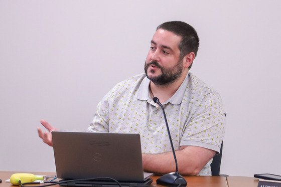 A gentleman sitting at a table with a laptop in front of him speaks into a microphone.
