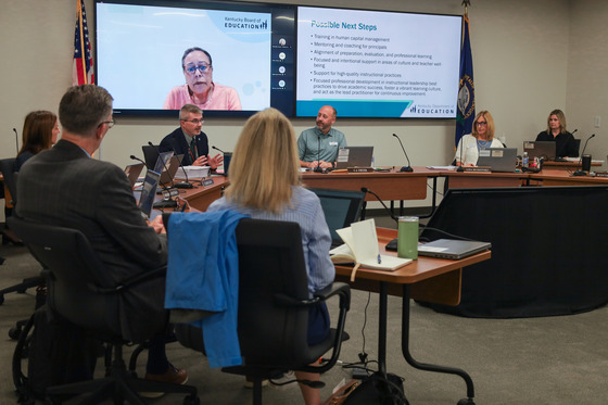 Robbie Fletcher sits in a board room with a few other people sitting around him and a screen behind him shows a woman