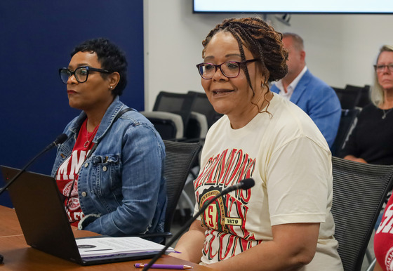 Two women sit at a table with microphones as the one closest to the camera speaks