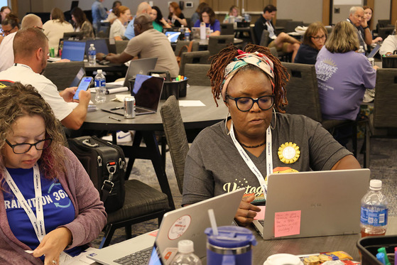 Two women sit at computers working, surrounded by tables full of teachers either working on their computers or talking.