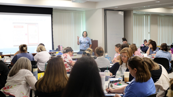 A group of teachers sit at tables, taking notes while listening to a speaker.