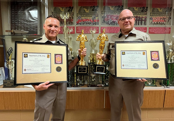Tom Krupp and Larry Lee pose in front of an awards case holding large framed certificates and statues. 