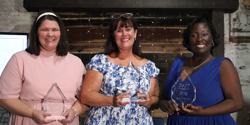 Three women pose for a photo together and smile while holding awards
