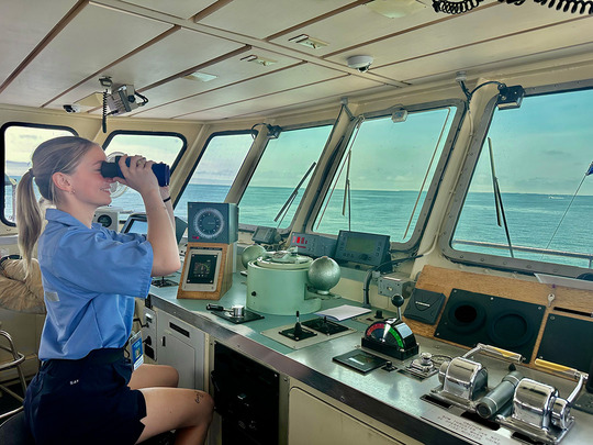 A woman in a blue uniform uses binoculars on a ship’s bridge, looking out to sea through large windows above navigation equipment.