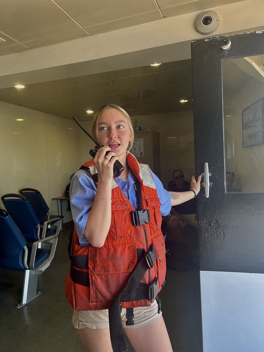 A woman wearing a red life vest and holding a walkie-talkie stands by a door on a boat, with seats and people visible in the background.