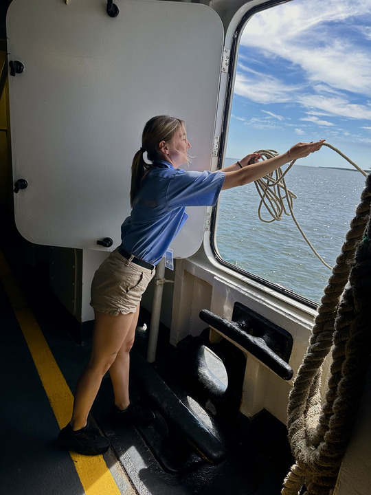 A person in shorts and a blue shirt throws a rope out of a ship’s doorway toward the water, under a partly cloudy sky.