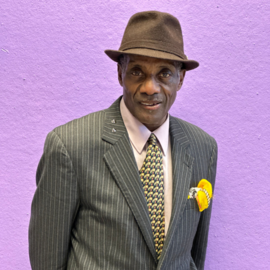 Laurence in a brown suit and tie and hat standing in front of a purple wall, smiling.