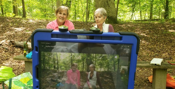 In the background are two women sitting on a bench in a wooded area and in the foreground is a tablet that is recording their educational lesson.