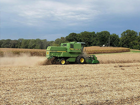 A green and yellow John Deere combine harvester moving from right to left across a harvested field under an overcast sky.