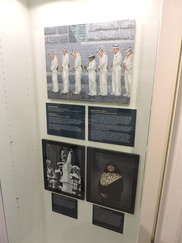 A  painting of seven midshipmen in white uniforms bowing their heads, with two framed photographs and biographical plaques below it.