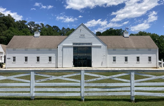 White clapboard dairy barn with a cross-gable roof, glass entry doors, and cupolas, seen behind a white wooden fence.