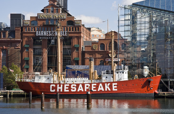 Red historic lightship named CHESAPEAKE docked in a city harbor, set against large brick and glass buildings.