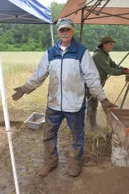 A man covered in mud stands smiling under a canopy at an outdoor archaeological dig site alongside screening equipment.