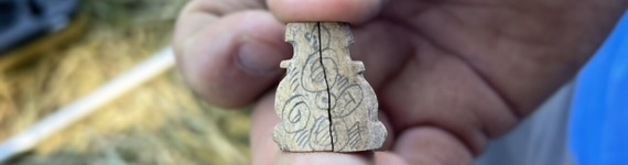 A close-up view of a person holding a small, intricately carved bone or wood artifact with spiral designs between fingers.