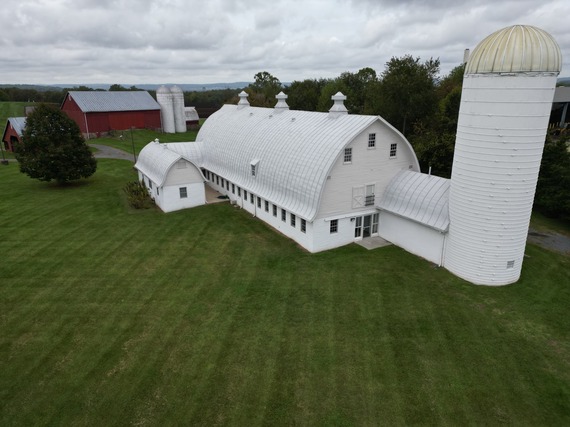 Aerial view of a large white barn with a curved roof, a tall white silo, and green lawn under an overcast sky.