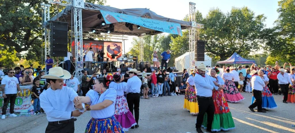 Dancers in traditional attire perform outdoors near a covered concert stage with a banner that reads "Nuestras Raices 2024".