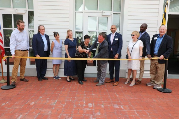 A group of people stands on a brick patio outside a white building for a ceremonial yellow ribbon-cutting.
