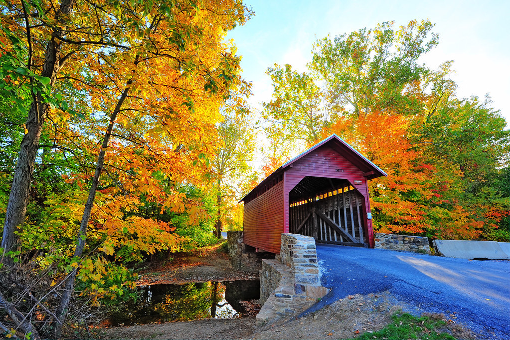 A historic red covered bridge spanning a stream, framed by vibrant yellow and orange autumn foliage.