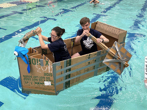 A man and woman paddle their cardboard boat across a pool