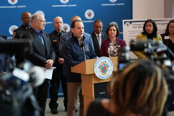 Councilmember Glass speaks at a press conference. Behind him stand other Councilmembers, the County Executive, and the Police Chief.