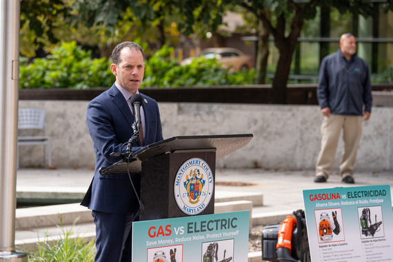 Councilmember Glass speaks at the bill signing in front of two posters explaining the difference between gas-powered and electric leafblowers