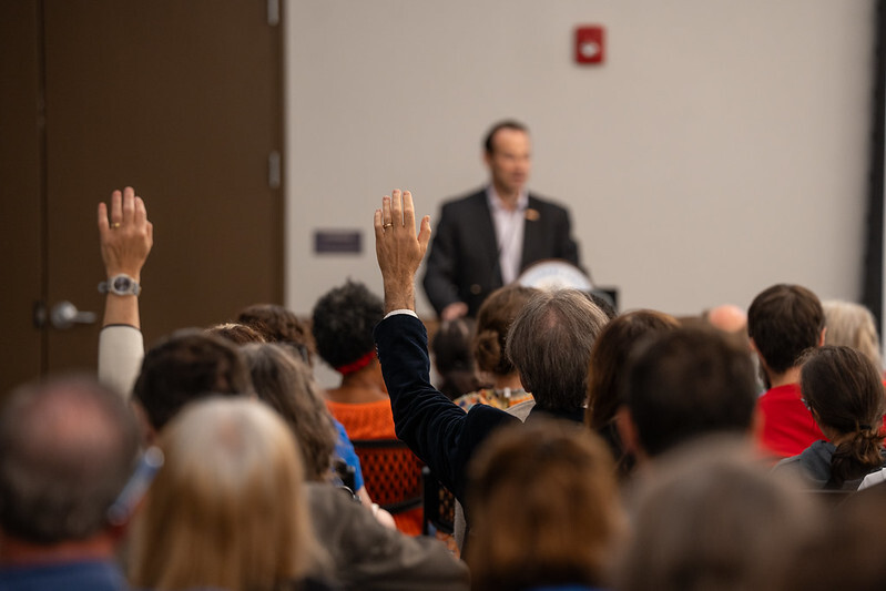 Two people raise their hands in a crowded audience with Council President Friedson standing at a podium.