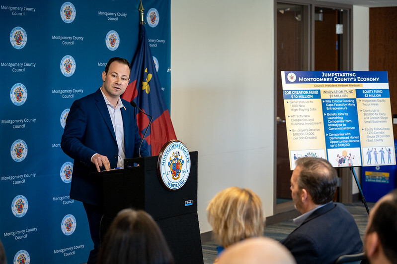 Council President Friedson speaks at a podium next to a poster reading “Jumpstarting Montgomery County’s Economy”.