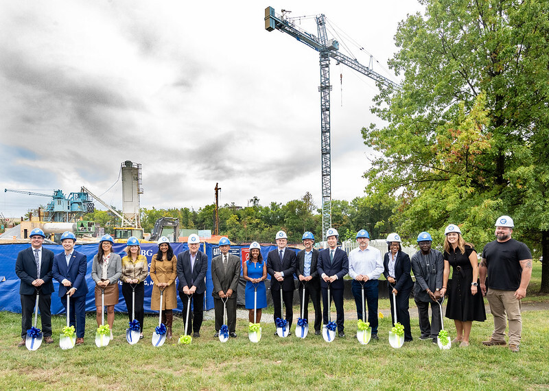 Group wearing hard hats and holding shovels with ribbons in front of a construction site for a groundbreaking event.