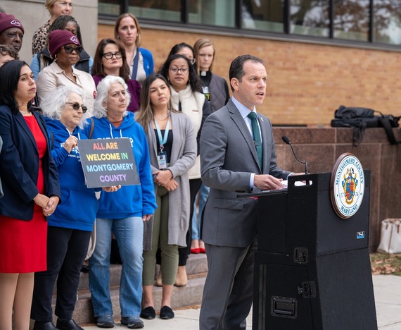 Councilmember Glass speaks at a podium in front of a crowd. A woman in the crowd holds a sign that reads "All are welcome in Montgomery County"