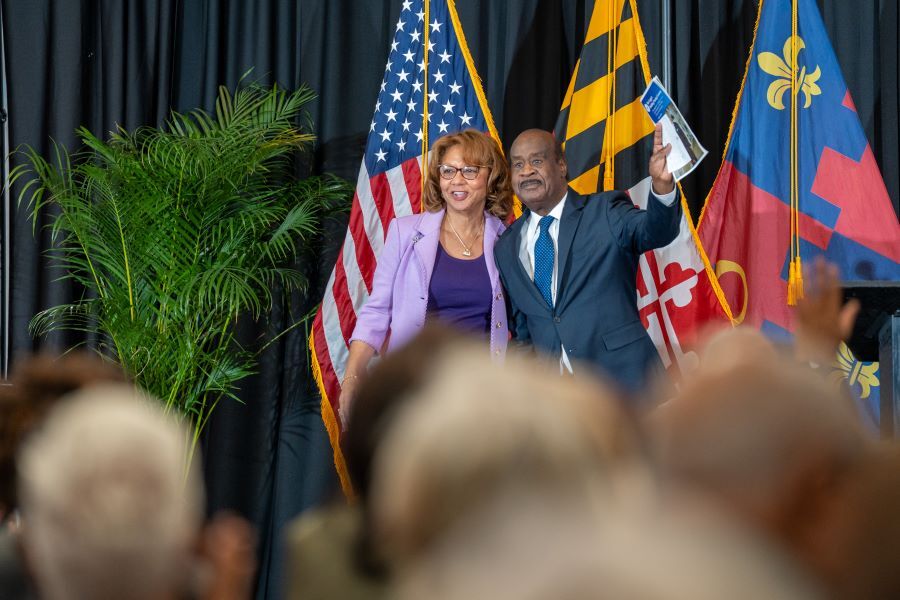 Catherine and Ike Leggett smile and wave on stage in front of flag display.