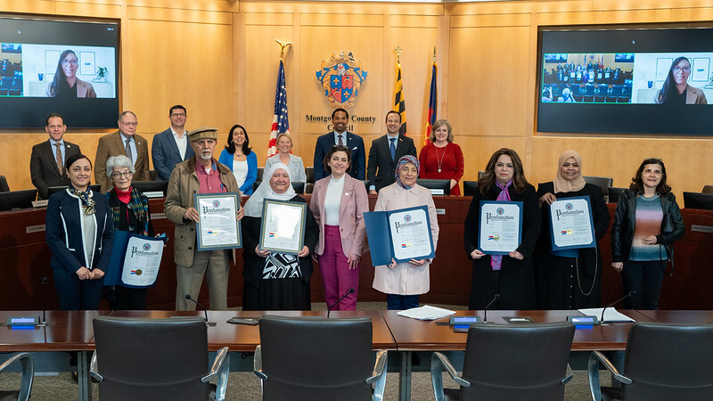 Councilmembers and Middle Eastern American residents stand for a group photo in the Council chamber holding proclamations.